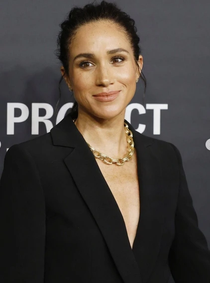 Meghan Markle wearing a black tailored pantsuit at a New York gala, no blouse, gold necklace, standing next to Prince Harry.